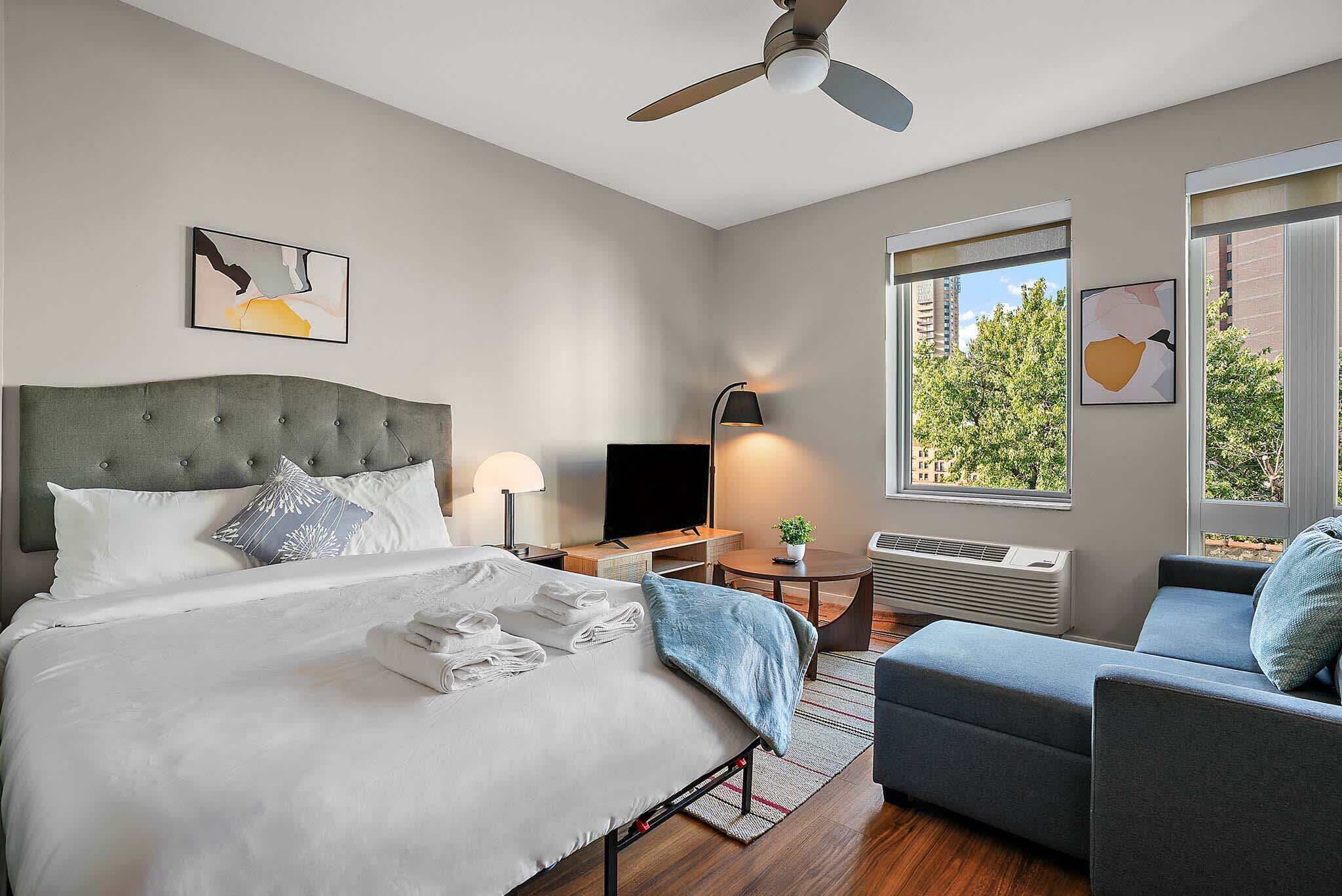 Camera d'hotel moderna con un letto grande, una scrivania, una sedia, una ventola a soffitto e ampie finestre vista alberi.