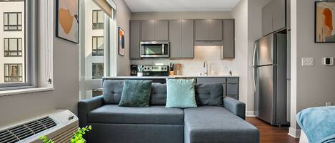 A modern living room with a sectional sofa, a dining table, and a kitchen area with grey cabinets and stainless steel appliances.