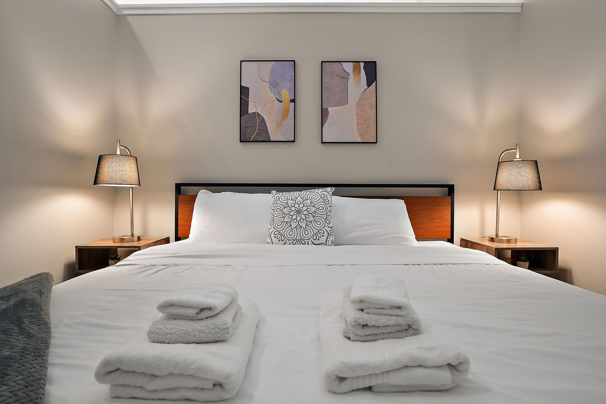 Una cama bien hecha con toallas dobladas, dos cuadros abstractos colgados en la pared y dos lámparas de noche.