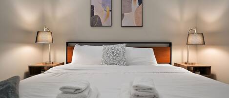 Una cama bien hecha con toallas dobladas, dos cuadros abstractos colgados en la pared y dos lámparas de noche.