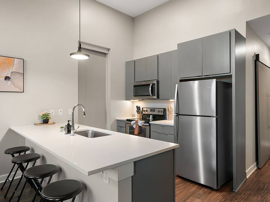A modern kitchen with grey cabinets, a stainless steel refrigerator, a microwave, and a white countertop. There are three black stools at the island.