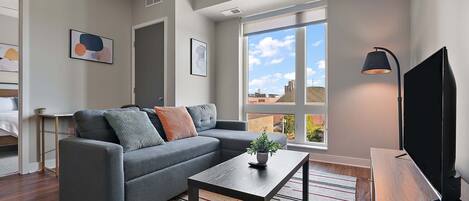 A modern living room with a grey sofa, a small table with a plant, a TV, and a large window with a city view.
