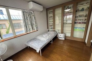 A small, single-bed room with a window, a fan, and a shelf with decorative items. - ShareHouse (Sapporo)