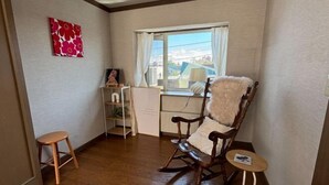 A room with a wooden floor, a rocking chair with a white cushion, a small wooden stool, a shelf with books and a plant, and a window with a view of buildings. - ShareHouse (Sapporo)