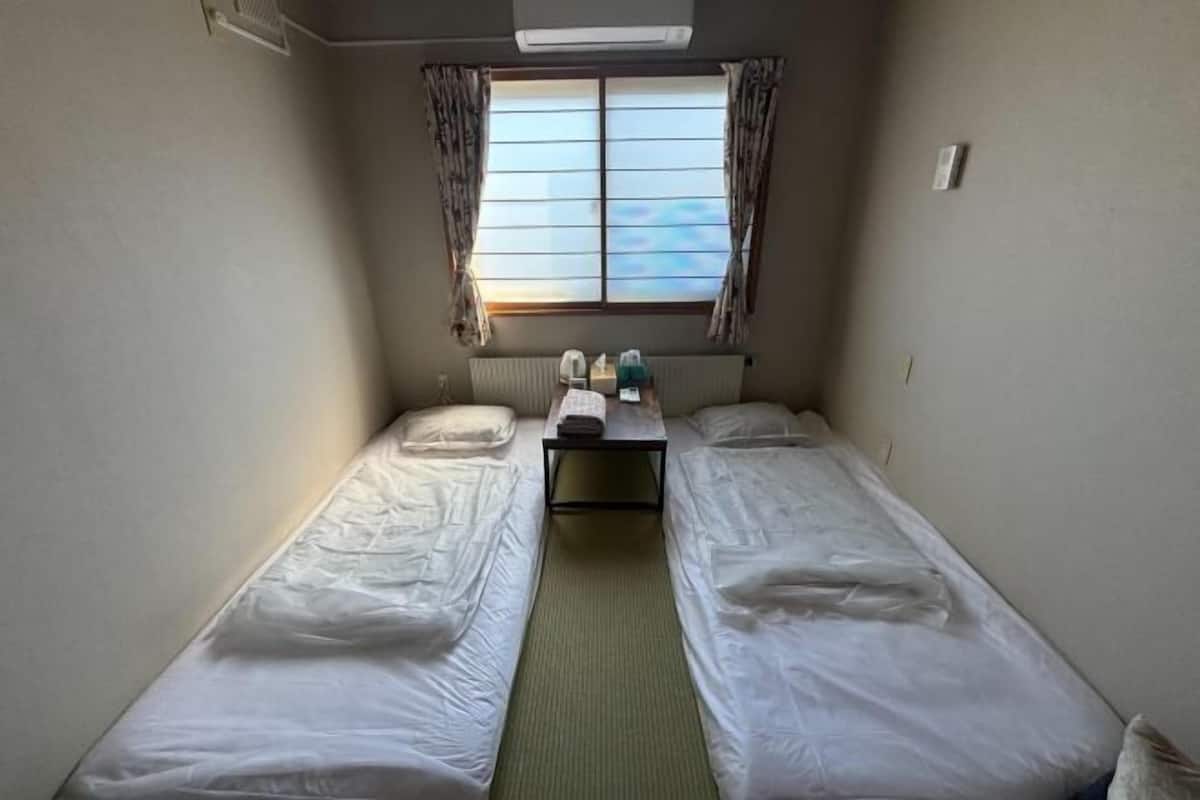 Une petite pièce avec deux lits simples, une table avec une chaise et une fenêtre avec des rideaux.