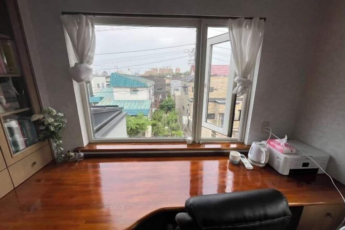 Une pièce avec une grande fenêtre, un bureau en bois, une chaise noire et une table de chevet avec une lampe.