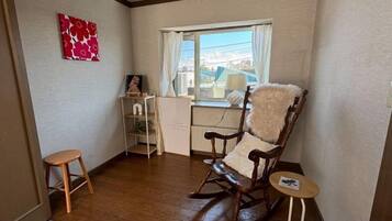 A room with a rocking chair, a small stool, a radiator, and a window with a view of buildings.
