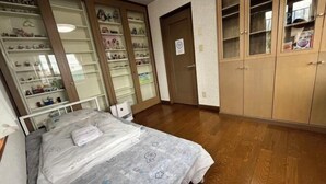 A bedroom with a bed, wooden wardrobe, and shelves with various items. - ShareHouse (Sapporo)