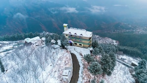 A multi-story building with a snow-covered roof, situated on a hillside with a river running in front. - Hilltop Resort, Narkanda (Theog)