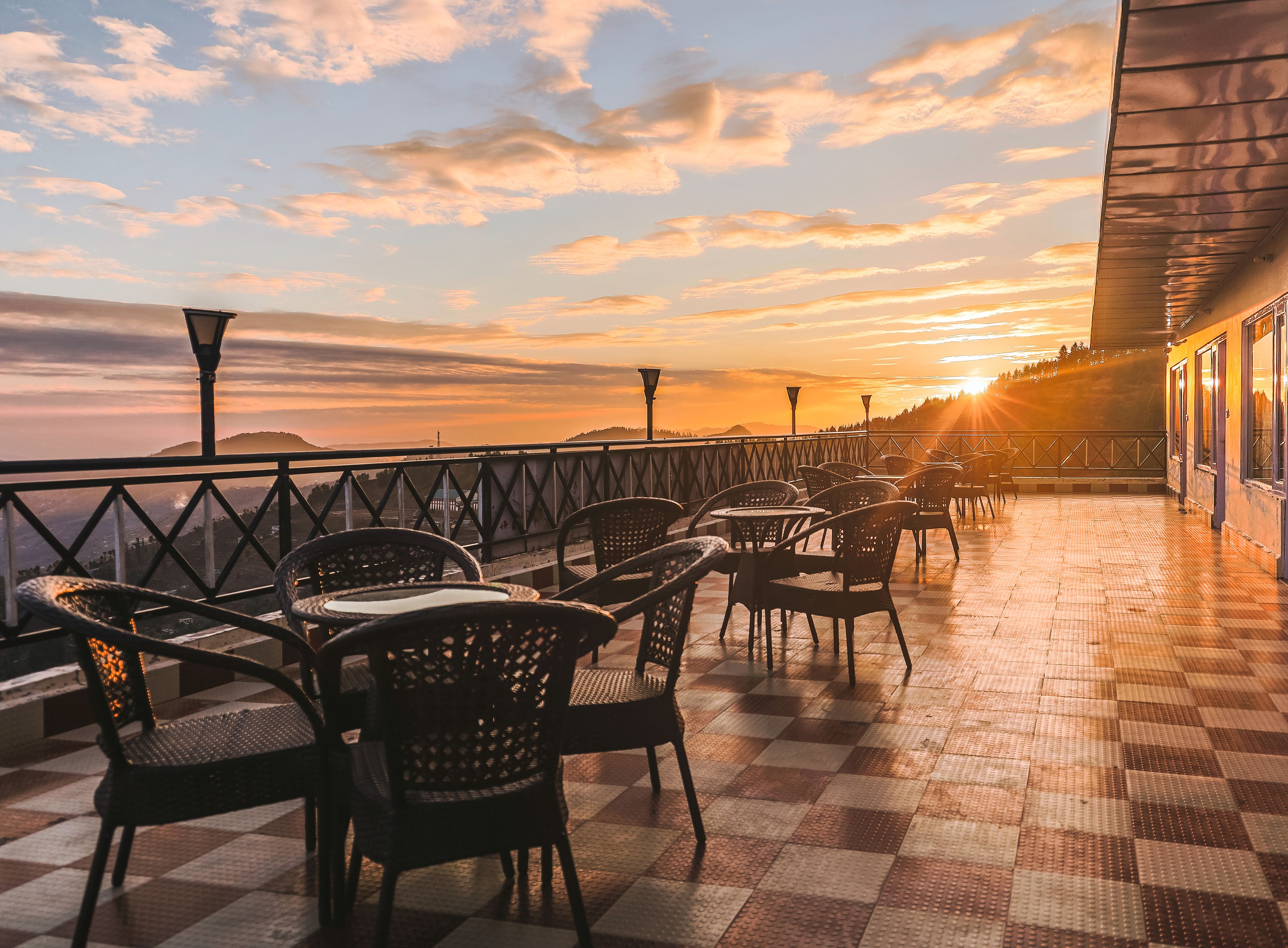 A terrace with checkered flooring, wicker chairs, and tables, overlooking a sunset.