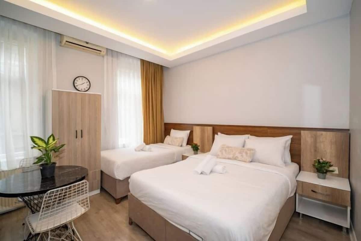 A hotel room with two beds, a small table, a chair, a clock, and an air conditioning unit.