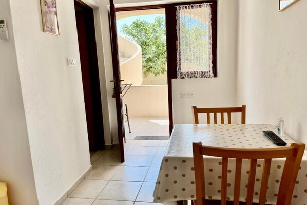 A dining area with a table and chairs, a window with lace curtains, and a view of the outside balcony. - Apartments Fortuna - Studio Apartment (Biograd na Moru)