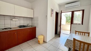 A compact kitchen with white cabinets, a sink, and a small trash bin. There is a dining area with a table and chairs, and a window with a curtain. - Apartments Fortuna - Studio Apartment (Biograd na Moru)