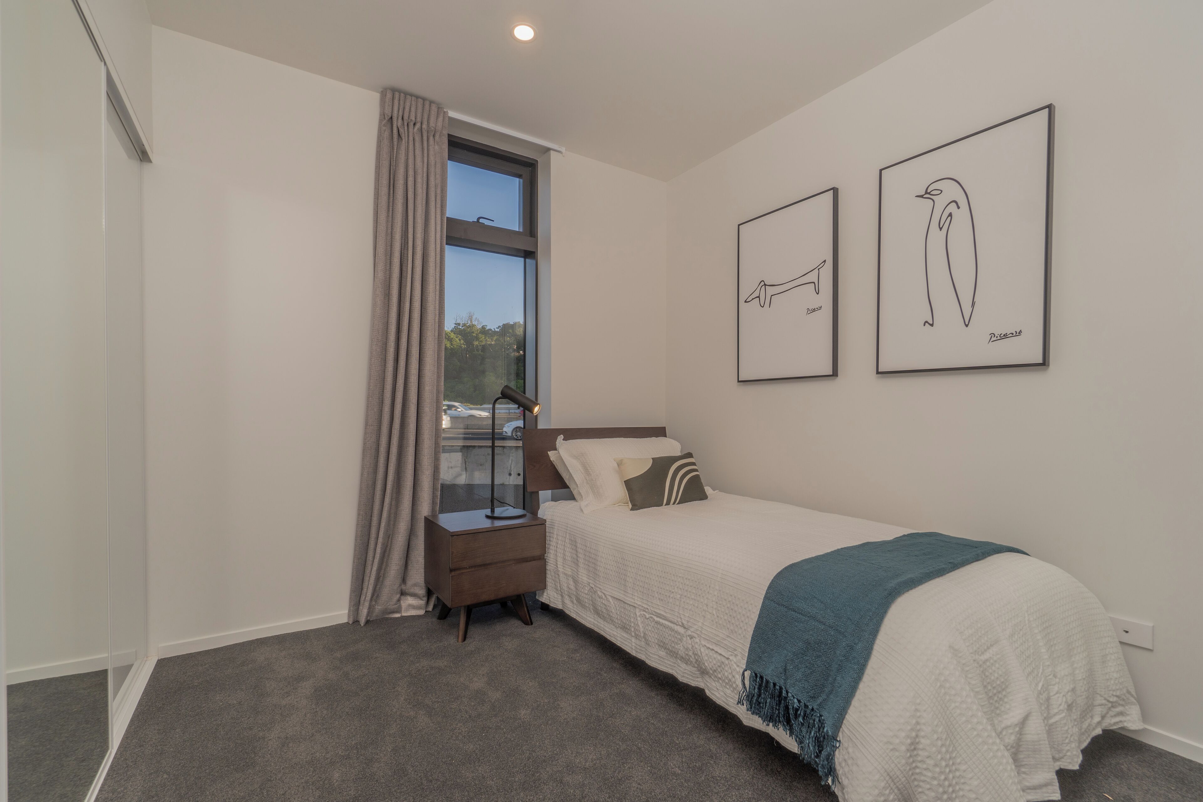 A bedroom with a bed, a nightstand, a window with curtains, and two framed pictures on the wall.
