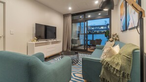 A modern living room with teal sofas, a flat-screen TV on a white cabinet, a coffee table, and a view of a balcony with a cityscape. - High End Centrally located apartment with carpark (Auckland)