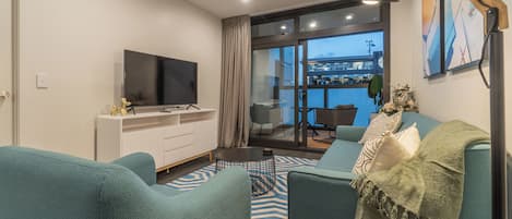 Un salon moderne avec des canapés de couleur turquoise, un téléviseur à écran plat posé sur un meuble blanc, une table basse et une vue sur un balcon donnant sur la ville.