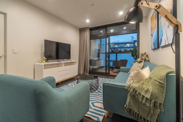 A modern living room with teal sofas, a flat-screen TV on a white cabinet, a coffee table, and a view of a balcony with a cityscape.