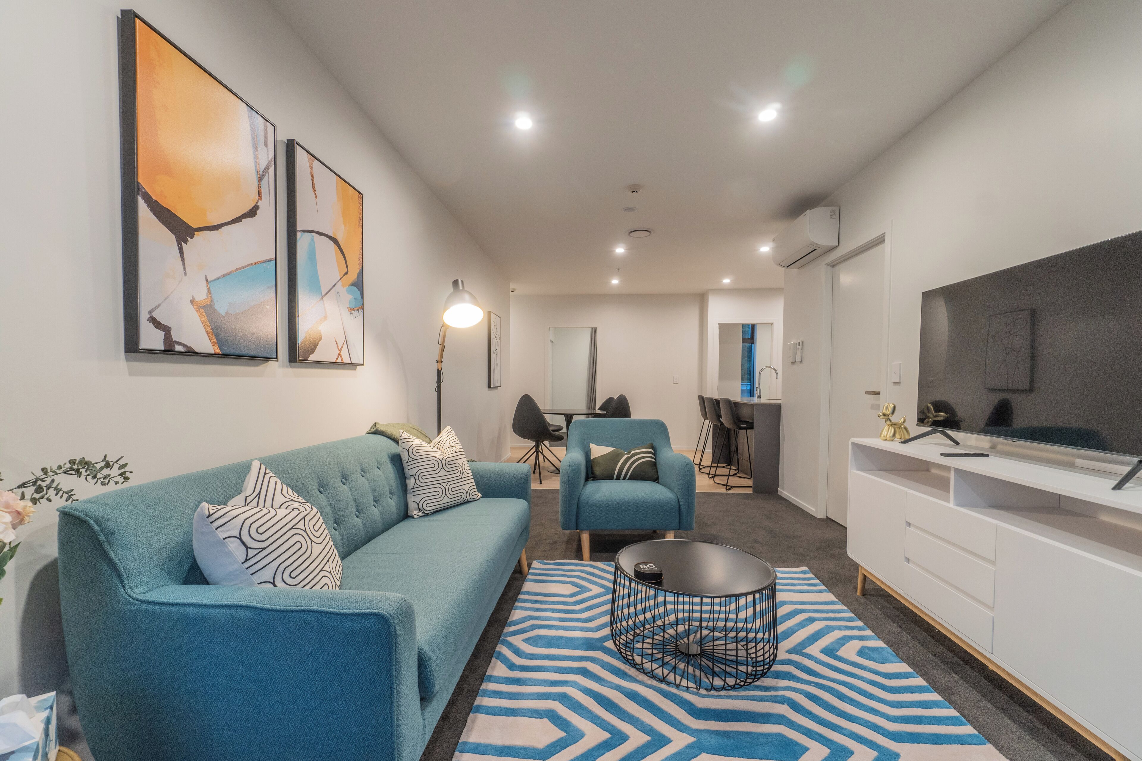 Un salon moderne avec un canapé de couleur turquoise, une table basse et un téléviseur posé sur un meuble blanc. Des tableaux abstraits ornent le mur, et on aperçoit un coin repas à l’arrière-plan.