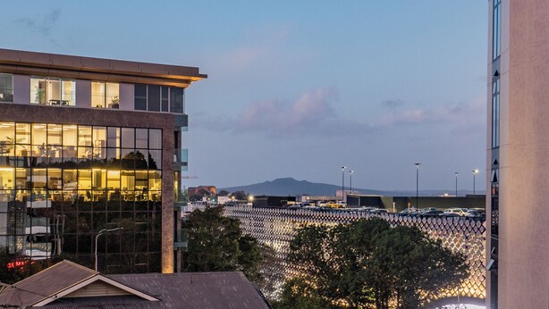 A modern building with large windows reflecting the interior lights, a parking area with cars, and a mountain in the background. - High End Centrally located apartment with carpark (Auckland)