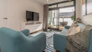 A modern living room with teal sofas, a flat-screen TV, a dining area with a glass table, and a view of a building through large windows. - High End Centrally located apartment with carpark (Auckland)