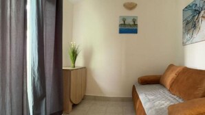 A room with a sofa, a small side table with a plant, a painting on the wall, and a curtain partially drawn. - One bedroom apartment with balcony-APP4 (Podaca)