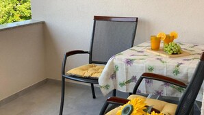 Un balcon avec deux fauteuils en osier, une table recouverte d’une nappe à motifs floraux et une petite table sur laquelle sont disposés des fruits et des boissons.