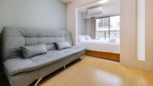 A modern hotel room with a grey sofa, a bed, and a large window with an air conditioning unit. - R Home Sakuragawa Riverside (Osaka)