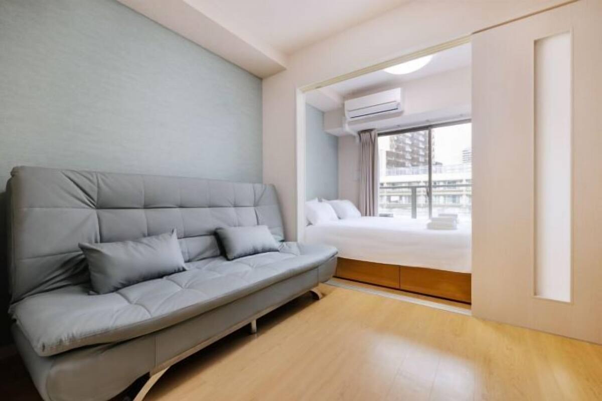 A modern hotel room with a grey sofa, a bed, and a large window with an air conditioning unit.