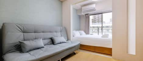 Una habitación de hotel moderna con un sofá gris, una cama y un ventanal con un equipo de aire acondicionado.