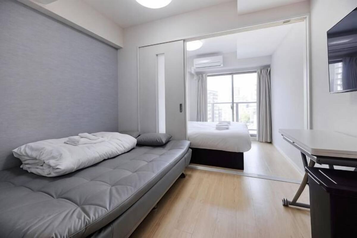 A modern, compact room with a grey sofa, a small desk, and a sliding door leading to a balcony.