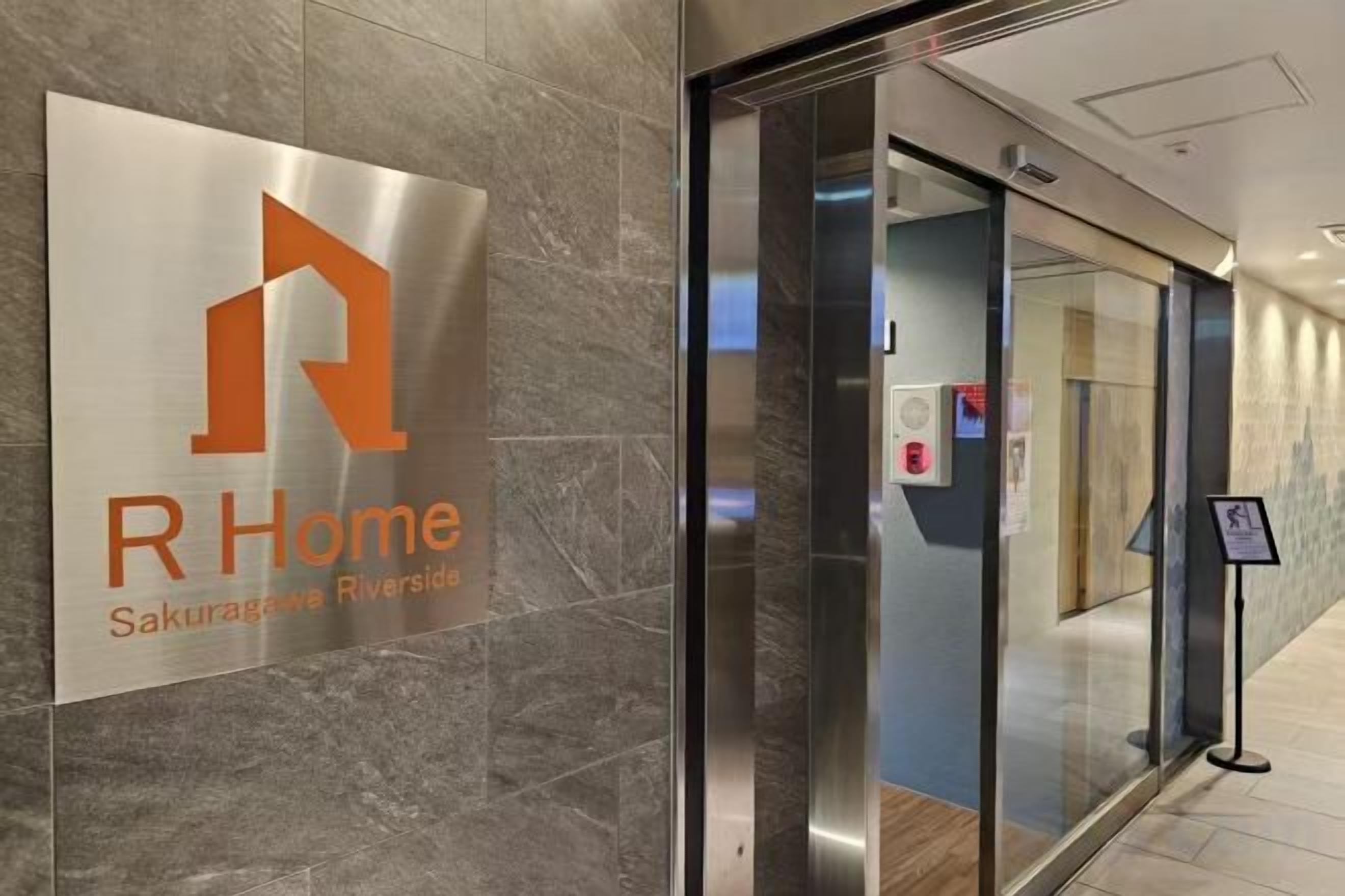 A modern hotel entrance with a glass door, a sign for RHome Sakuragawa Riverside, and a fire alarm panel.