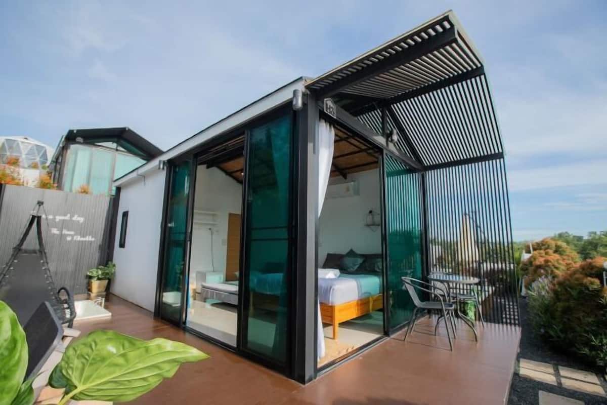 Ein modernes, kompaktes Wohnambiente mit einem Schlafzimmer mit Glasfassade, einer Terrasse mit Tisch und Stühlen sowie einer Hängematte.