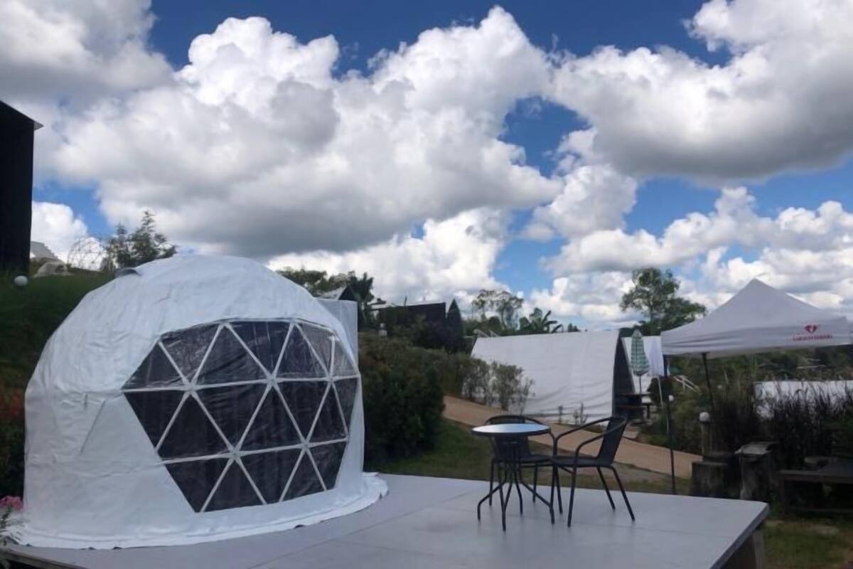 Eine kuppelförmige Struktur mit geometrischem Fensterbild, ein Tisch mit Stühlen und ein Zelt im Hintergrund.