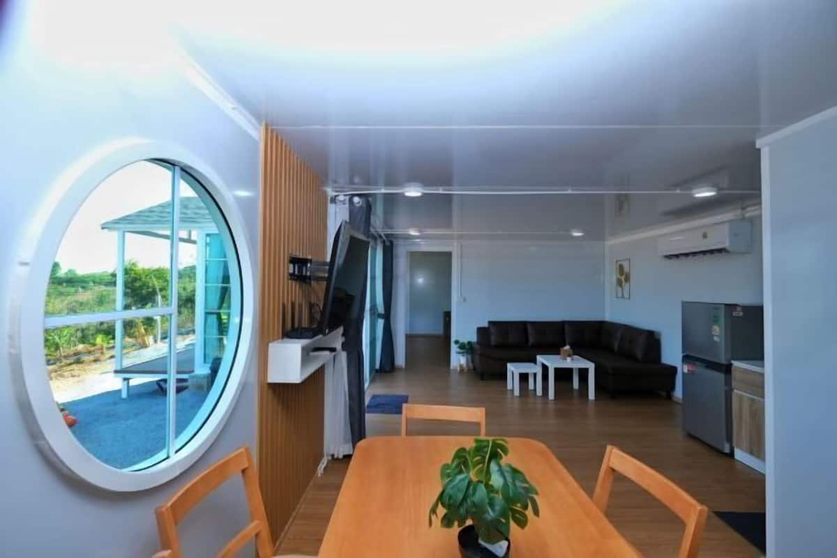 Ein modernes Wohnzimmer mit rundem Fenster, einem Esstisch mit Stühlen, einem schwarzen Sofa, einem kleinen Couchtisch und einer Küchenzeile inklusive Kühlschrank.