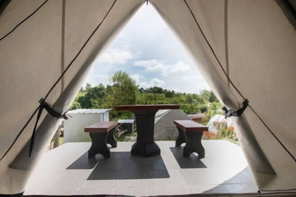 Ein Zelt mit Tisch und zwei Stühlen vor einer Kulisse aus Grünflächen und Gebäuden.