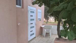 A small outdoor area with a white plastic chair, a white door, and a tree with large green leaves. - Apartments Rubbi - Studio with Terrace (Vrsi)