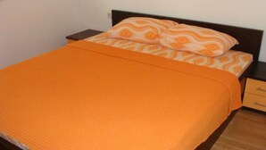 Een eenpersoonsbed met een oranje sprei en passende kussens, een nachtkastje van hout met lades, en een effen muur.
