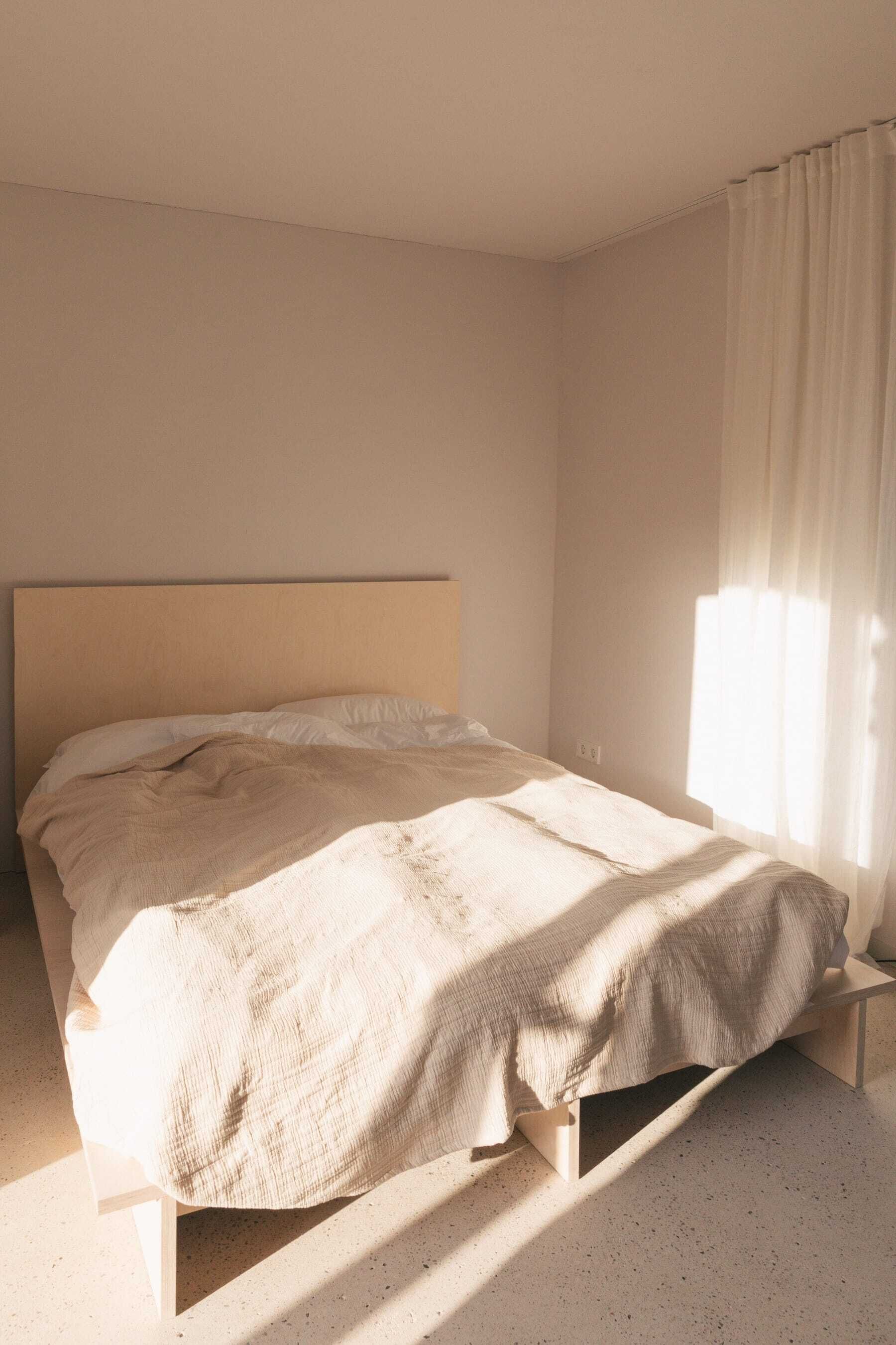 Un letto con struttura in legno e materasso beige in una stanza dalle pareti spoglie e una finestra con tende leggere.