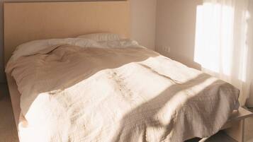 Un letto con struttura in legno e materasso beige in una stanza dalle pareti spoglie e una finestra con tende leggere.