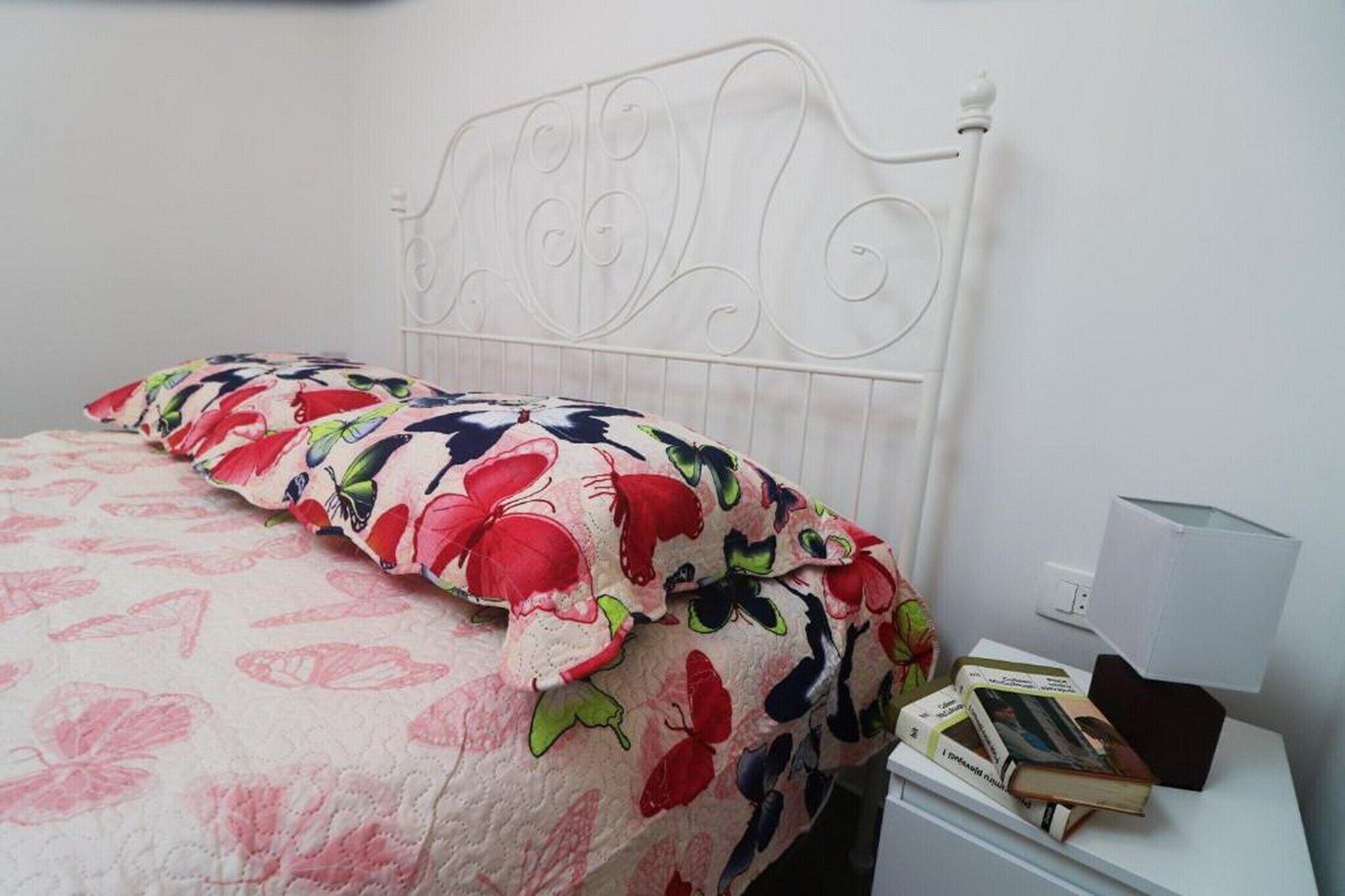A bed with a floral-patterned bedspread, a white metal headboard, and a bedside table with books and a lamp.