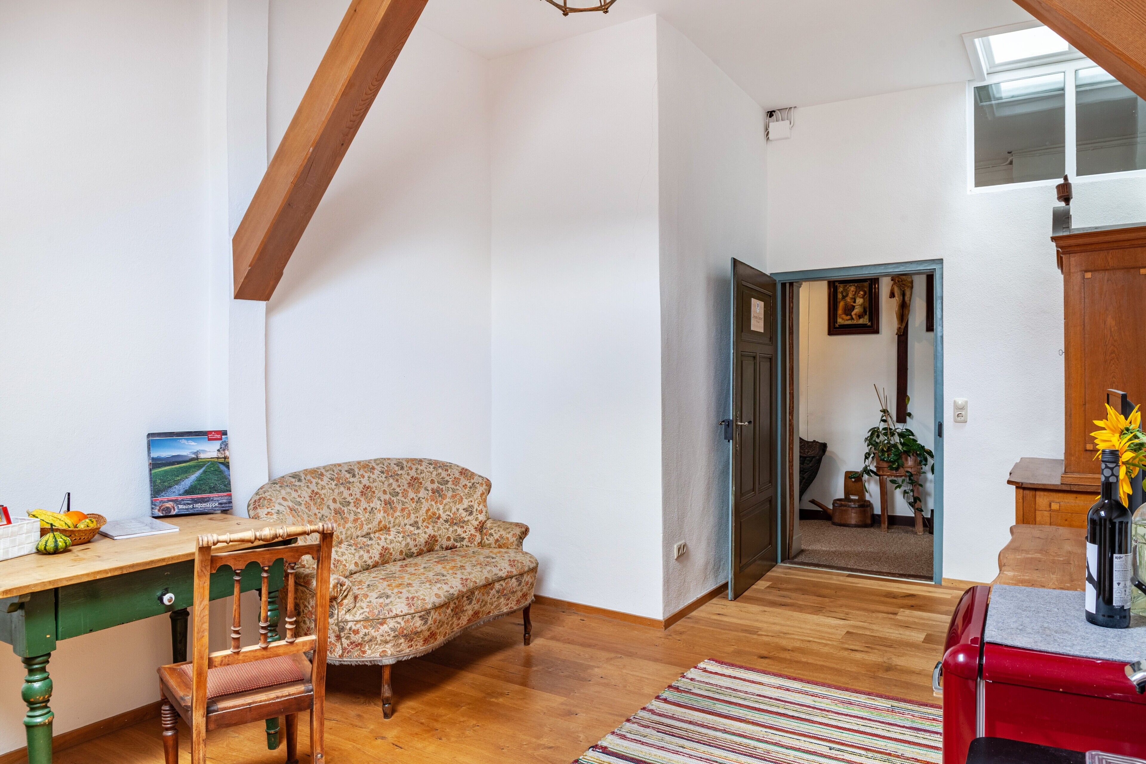 A room with a wooden desk, a floral sofa, a red cabinet, and a view of a garden through an open door.