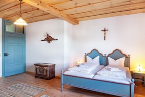Un dormitorio con techo de madera, puerta azul, cama con ropa blanca, mesita de noche y una cruz colgada en la pared.