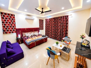 A modern hotel room with a red bed, a purple sofa, a dining table with chairs, and a ceiling fan. - Regalia Home (Jammu)