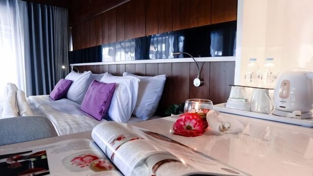 A hotel room with a bed, a table with a magazine, a red flower, and a coffee maker.