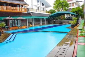 En swimmingpool med ett klart, blått vatten, omgiven av ett trädäck och ett avskärmat sittområde med parasoller.
