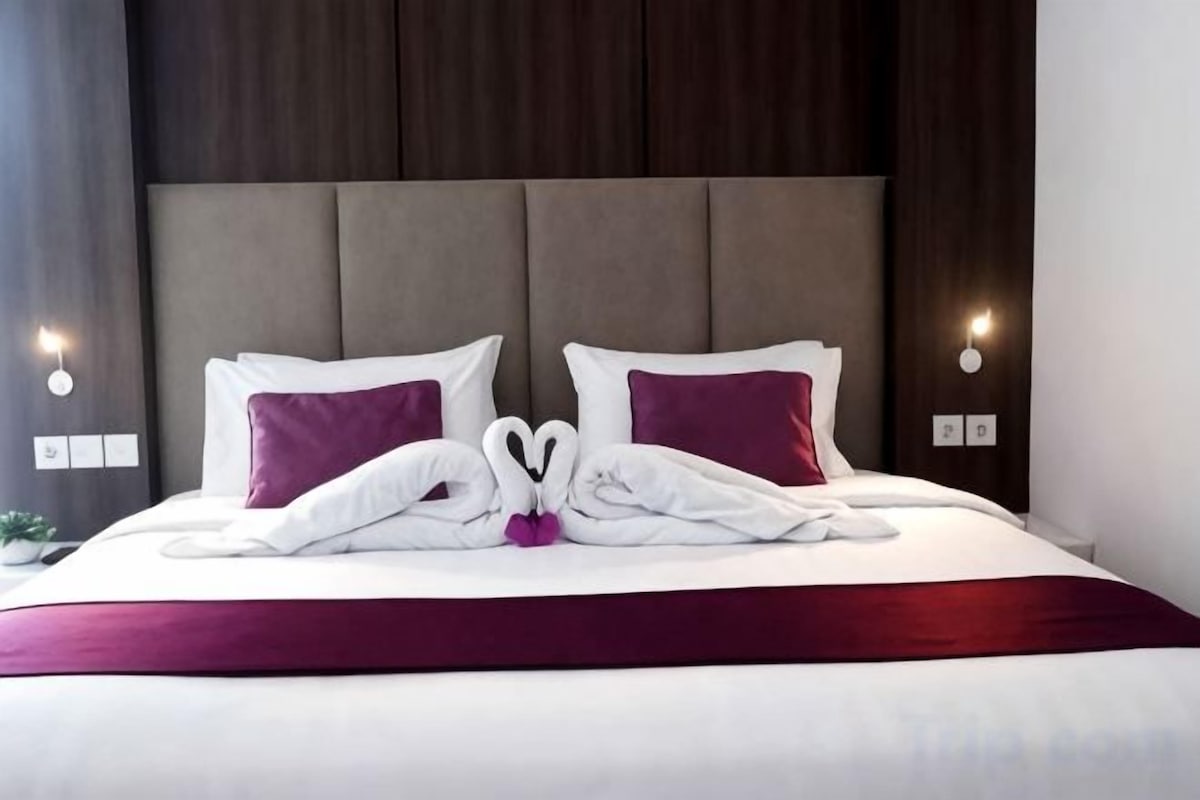Een keurig opgemaakt bed met een rode sprei, twee kussens en een handdoekje gevouwen in de vorm van een hart.