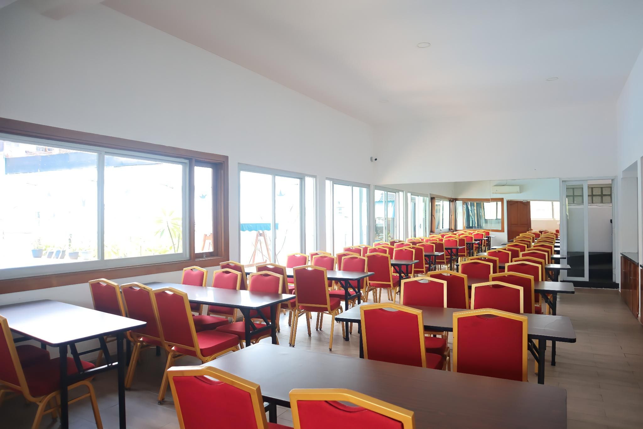 A large, well-lit room with rows of tables and chairs, large windows, and a door leading to another area.
