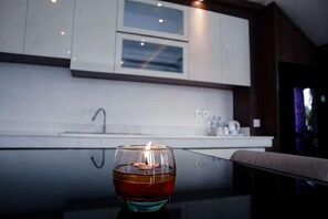 Una vela en un soporte de vidrio sobre una superficie reflectante en una cocina moderna con armarios blancos y un ventanal.