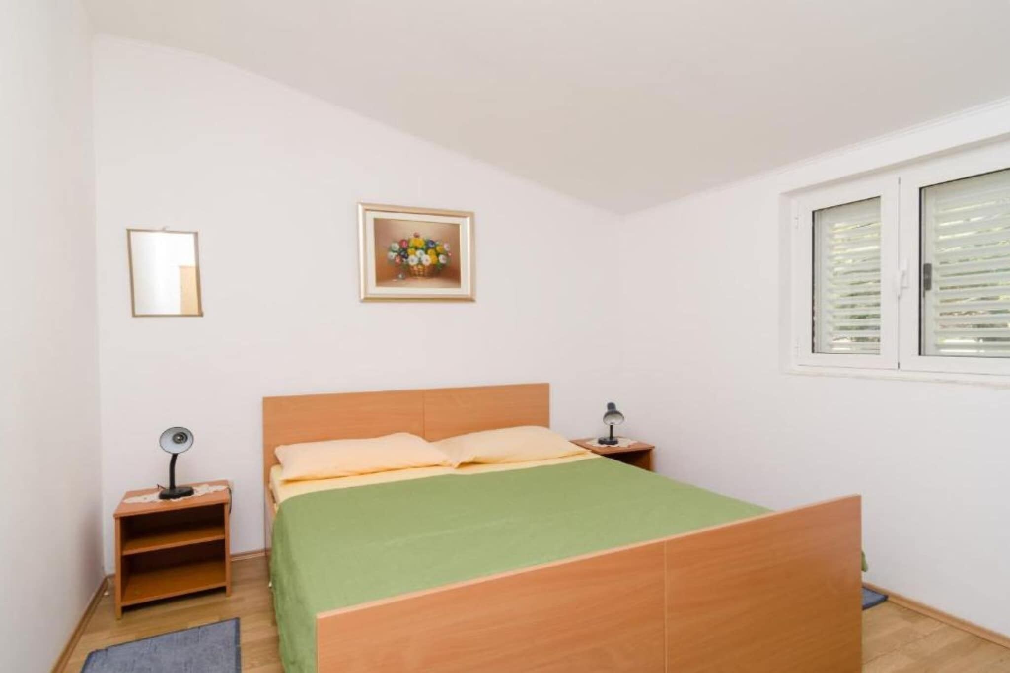 Une chambre avec un lit simple, une tête de lit en bois, une couvre-lit verte, deux tables de chevet avec des lampes, un tableau encadré au mur et une fenêtre avec des stores.