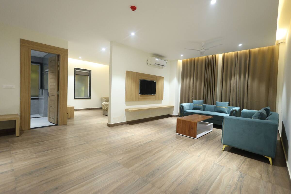 A spacious living room with a wooden floor, a blue sofa, a wooden coffee table, a mounted TV, and a ceiling fan.